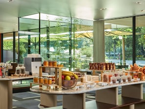 A breakfast buffet with various food items and condiments, set against a backdrop of large windows overlooking a green outdoor area.
