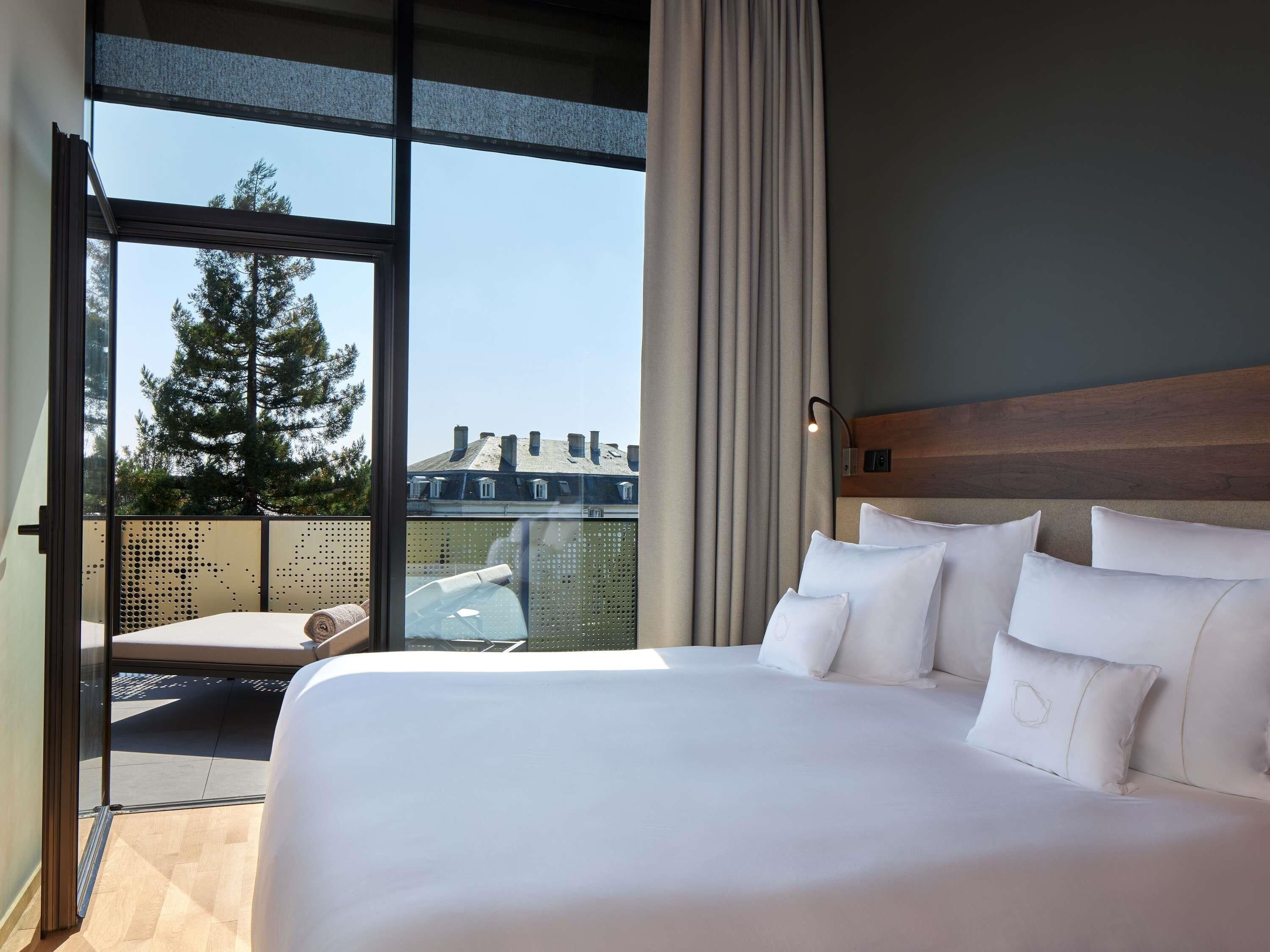 A hotel room with a large bed, a view of trees and buildings, and a balcony with a chair.