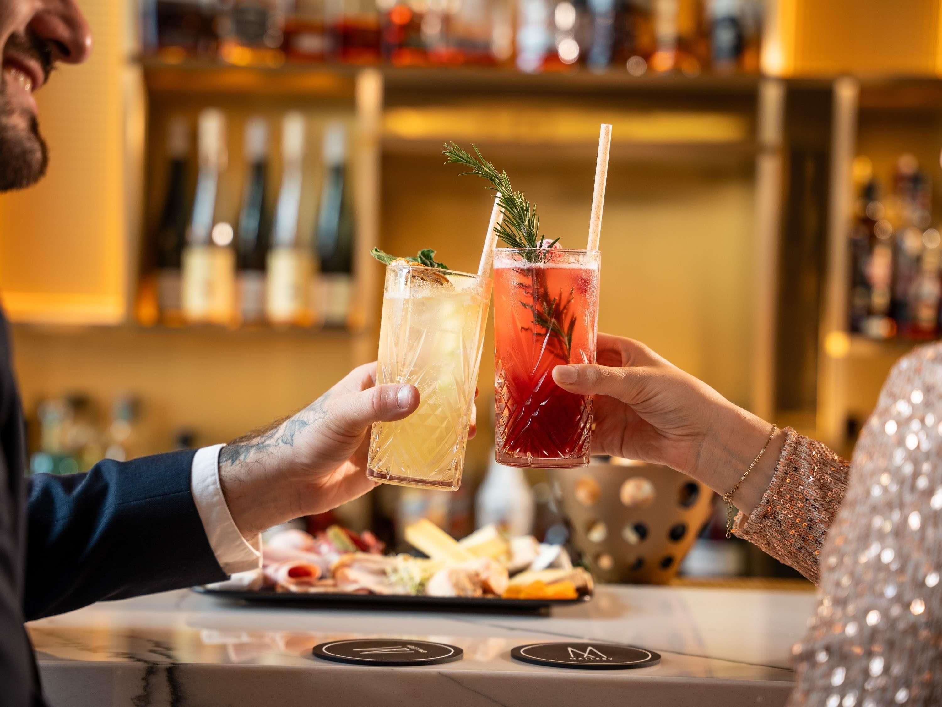 Dos personas brindando con bebidas en un bar, un menú y una barra con un logotipo.