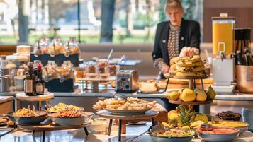 A buffet setup with various food items and beverages, a person serving, and a view of trees outside.