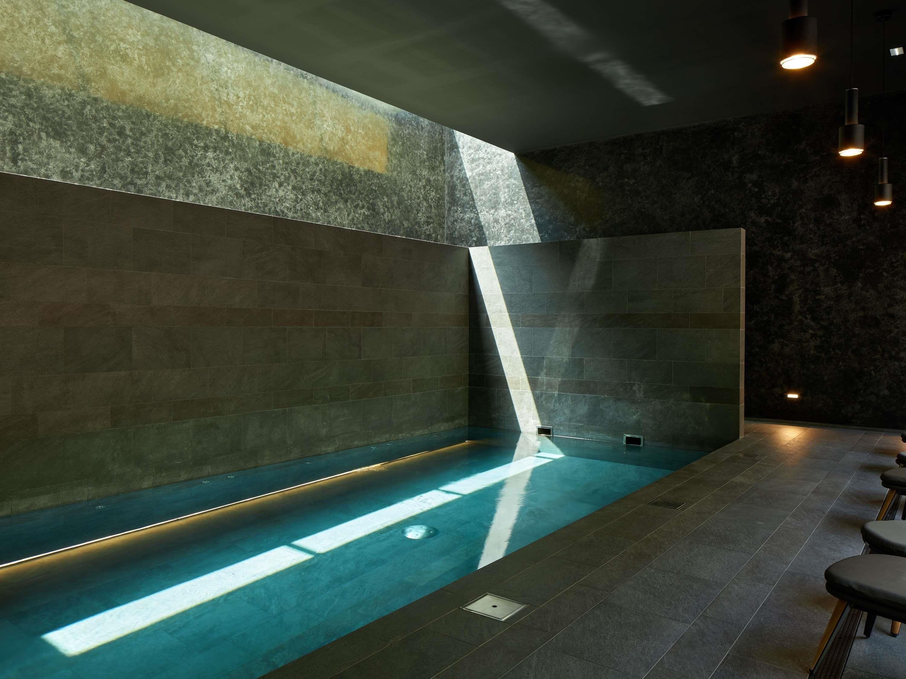 A modern indoor pool with a sloped ceiling, dark stone walls, and a seating area with bar stools.
