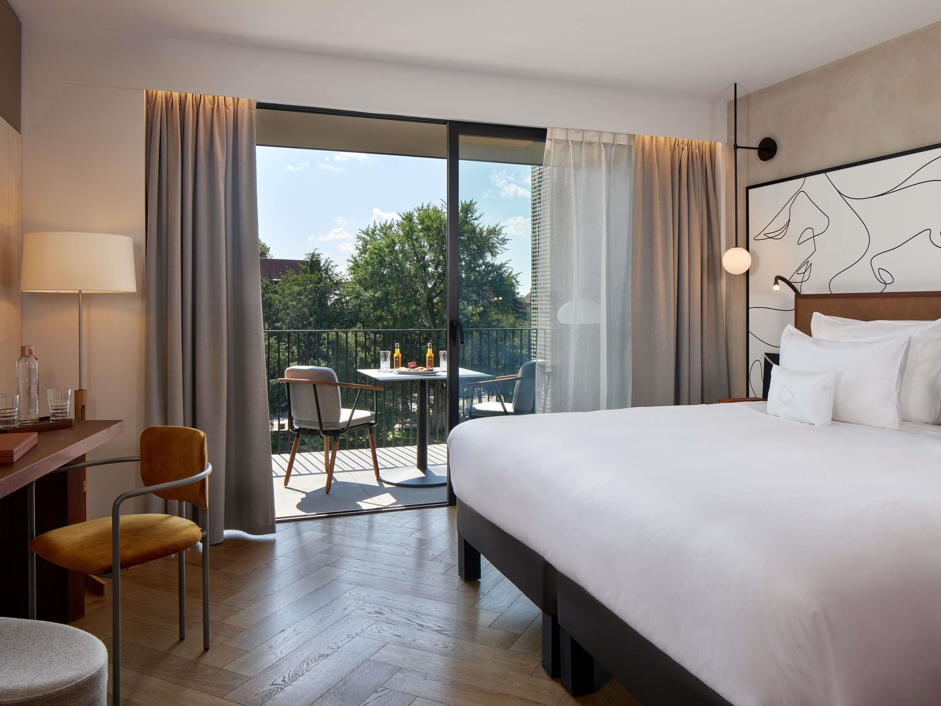 A hotel room with a bed, a desk with a chair, a table with chairs outside, and a balcony with curtains.