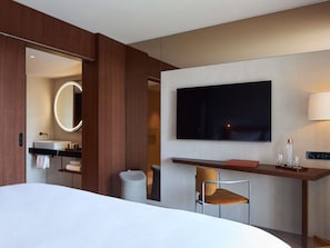 A hotel room with a bed, a desk with a chair, a TV, a mirror, and a bathroom with a sink and towel rack. - L'Esquisse Hotel & Spa Colmar - MGallery Collection (Colmar)