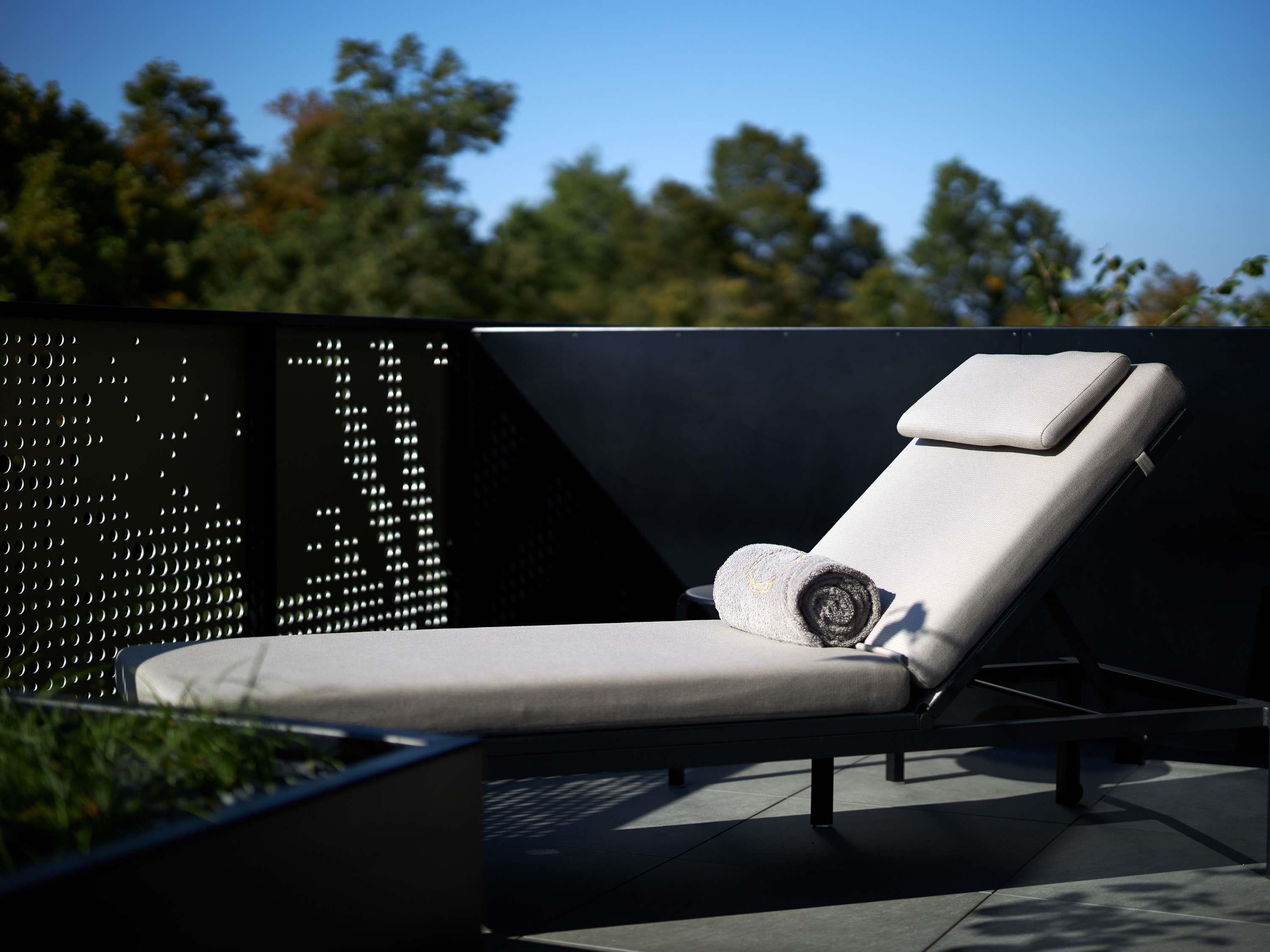 Ein moderner Lounge-Sessel mit Kissen, platziert auf einer Dachterrasse mit Metallzaun und Grünpflanzen im Hintergrund.
