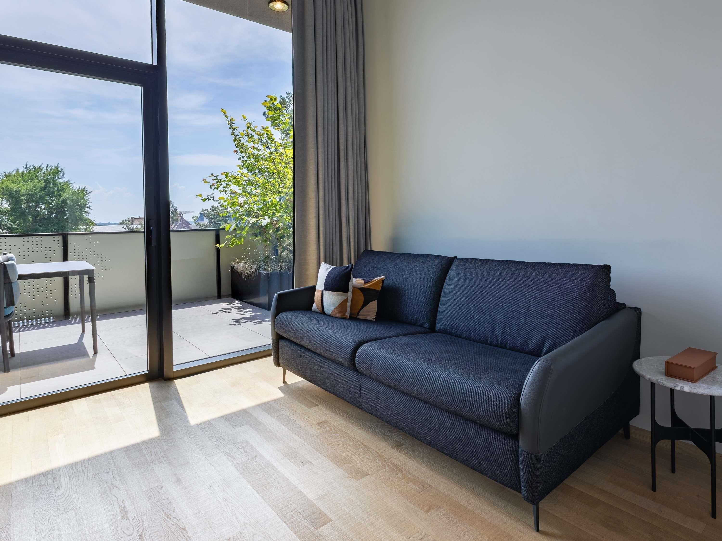 A modern living room with a sofa, a small table, and a balcony with a view of trees and buildings.