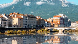 A historic town with a river reflecting buildings, a bridge, and a mountain in the background. - Hôtel Inn Design Grenoble Eybens Parc des Exposition (Eybens)