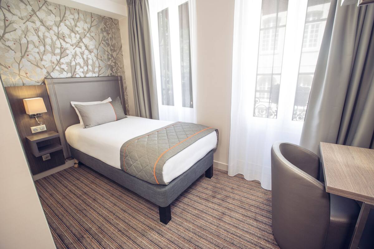 A hotel room with a bed, a desk, a chair, a nightstand with a lamp, and a window with curtains.