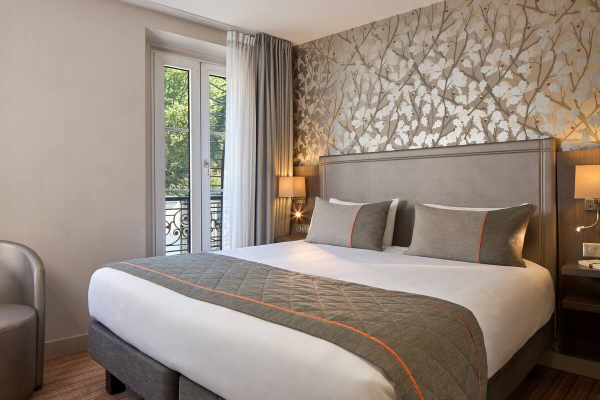 A hotel room with a large bed, a chair, a nightstand with a lamp, and a view of greenery through the window.