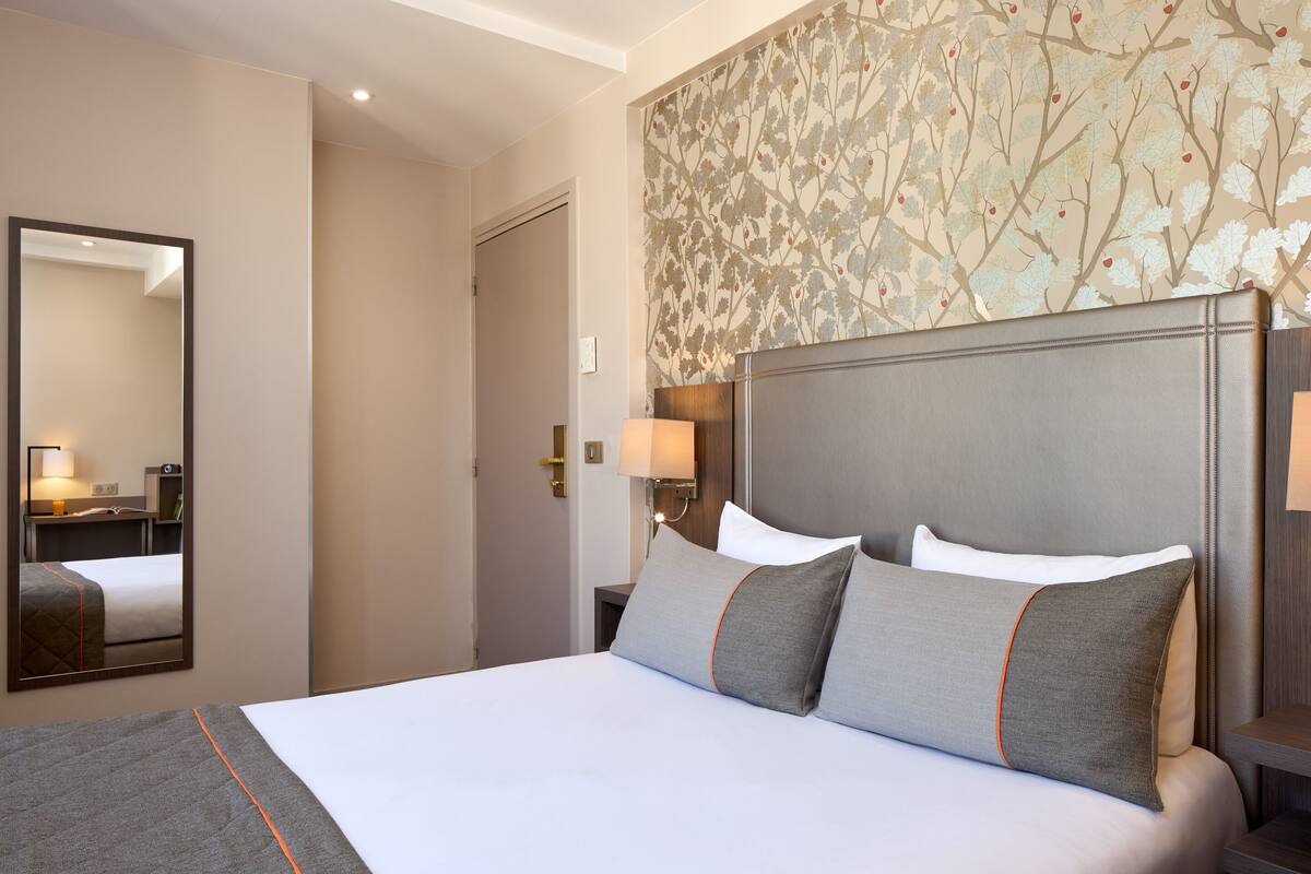 A hotel room with a large bed, a nightstand with a lamp, a mirror, and a door leading to another room.
