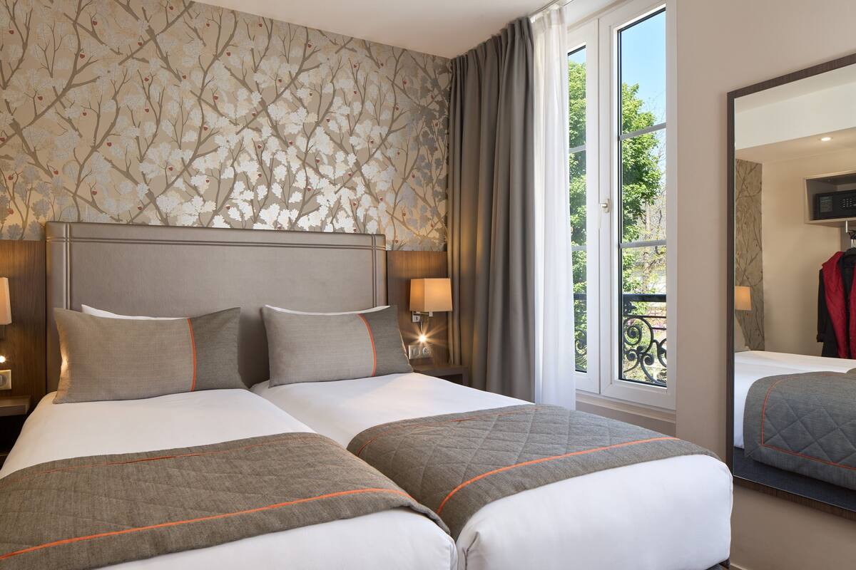 A hotel room with two beds, a large window with a view of greenery, and a mirror on the wall.