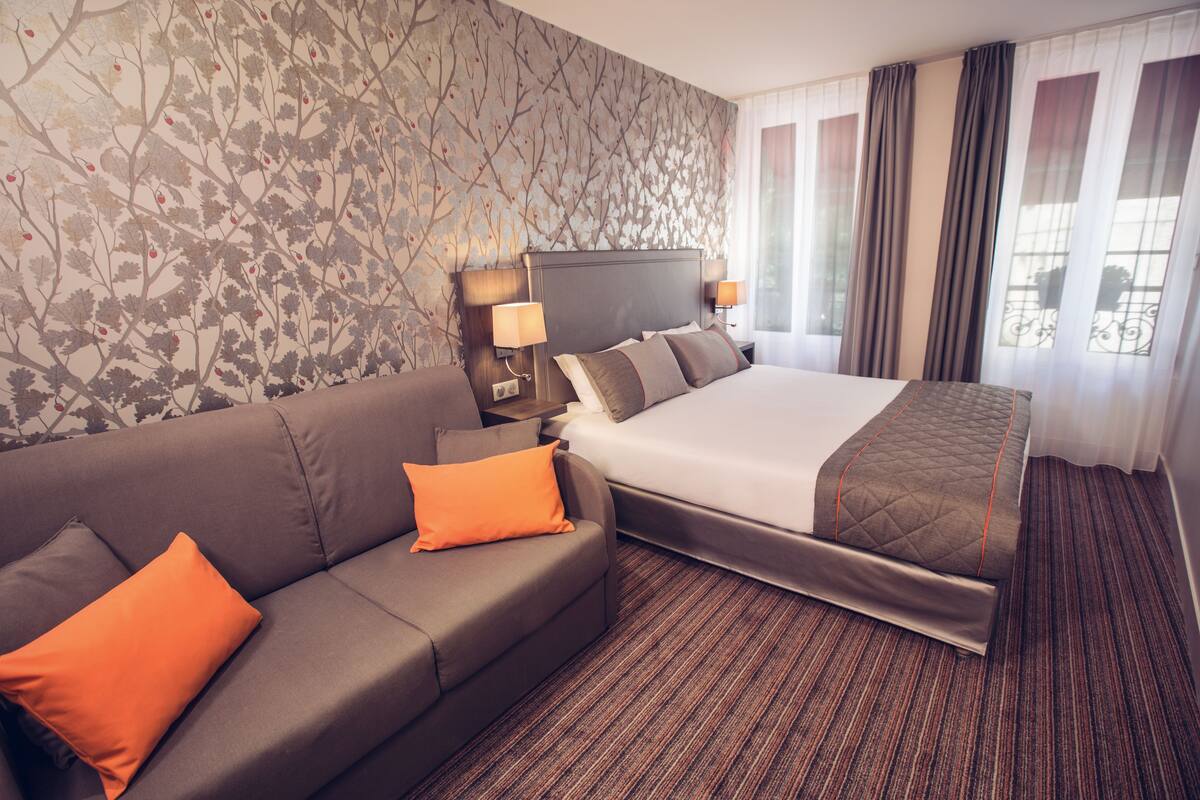 A hotel room with a bed, a sofa, a nightstand with lamps, and a patterned wallpaper.