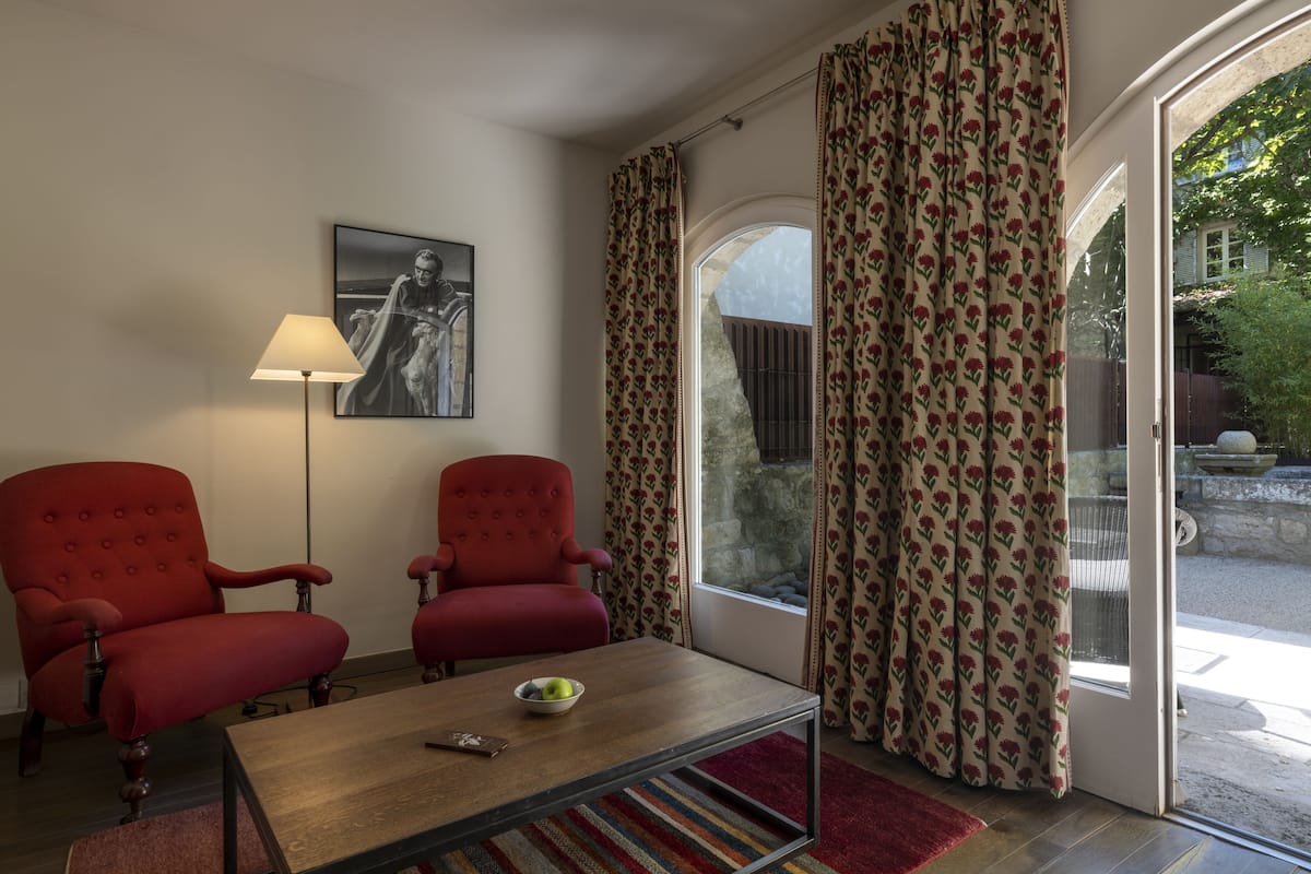 A room with two red armchairs, a wooden coffee table with a bowl and remote, a lamp, and a large window with floral curtains.