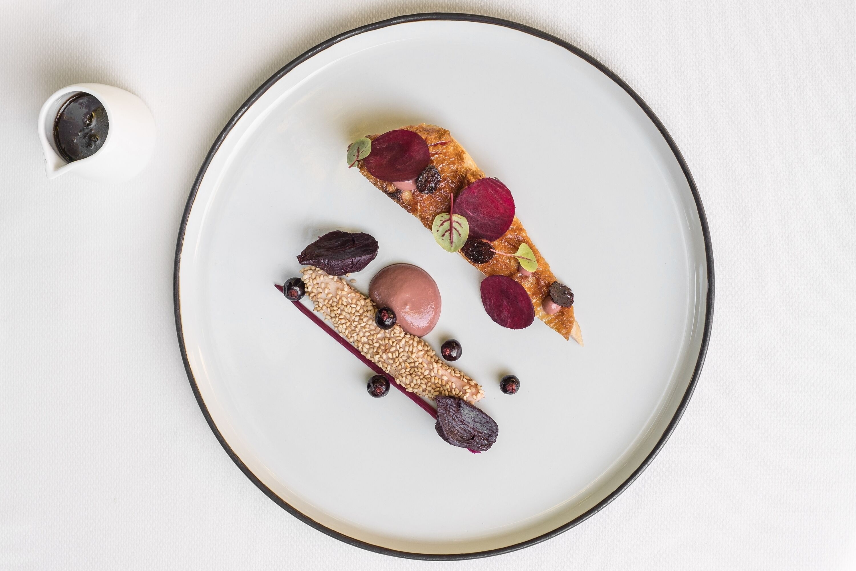 Un piatto bianco con un dessert composto da una fetta di torta guarnita con diversi frutti di bosco e semi, e una piccola tazza bianca con un oggetto scuro.