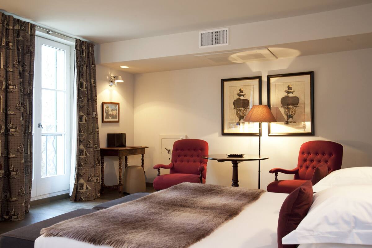 A hotel room with a bed, two red armchairs, a small table, a lamp, and a window with curtains.