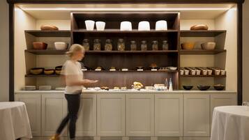 A buffet area with multiple shelves of dishes and jars, a person walking past, and tables with white tablecloths.