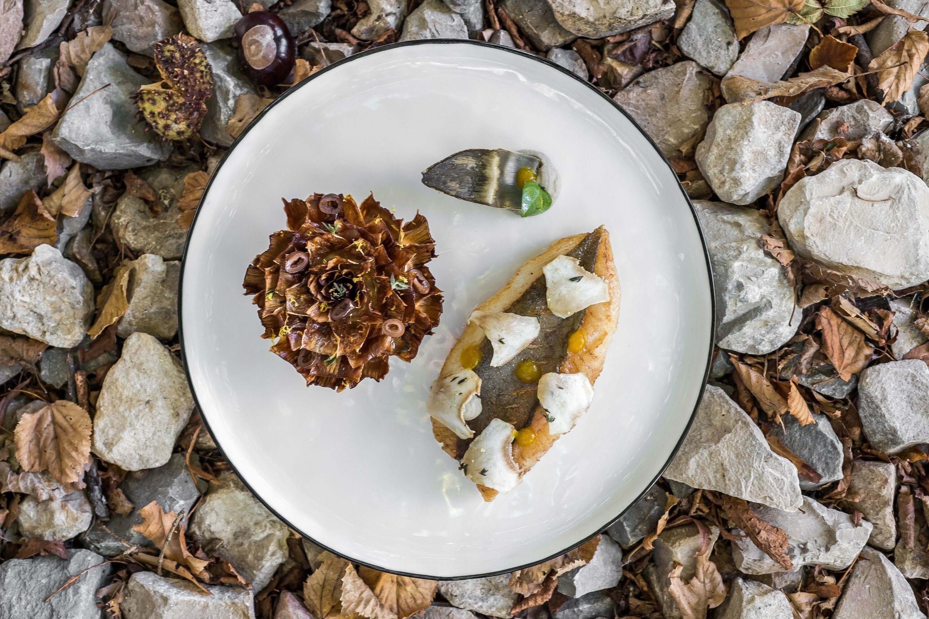 A white plate with a decorative item and two pieces of food on a bed of rocks and leaves.