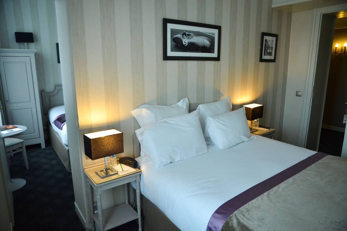 Una habitación de hotel con una cama grande, dos mesitas de noche con lámparas, un escritorio pequeño con una silla, un armario y un cuadro en la pared.