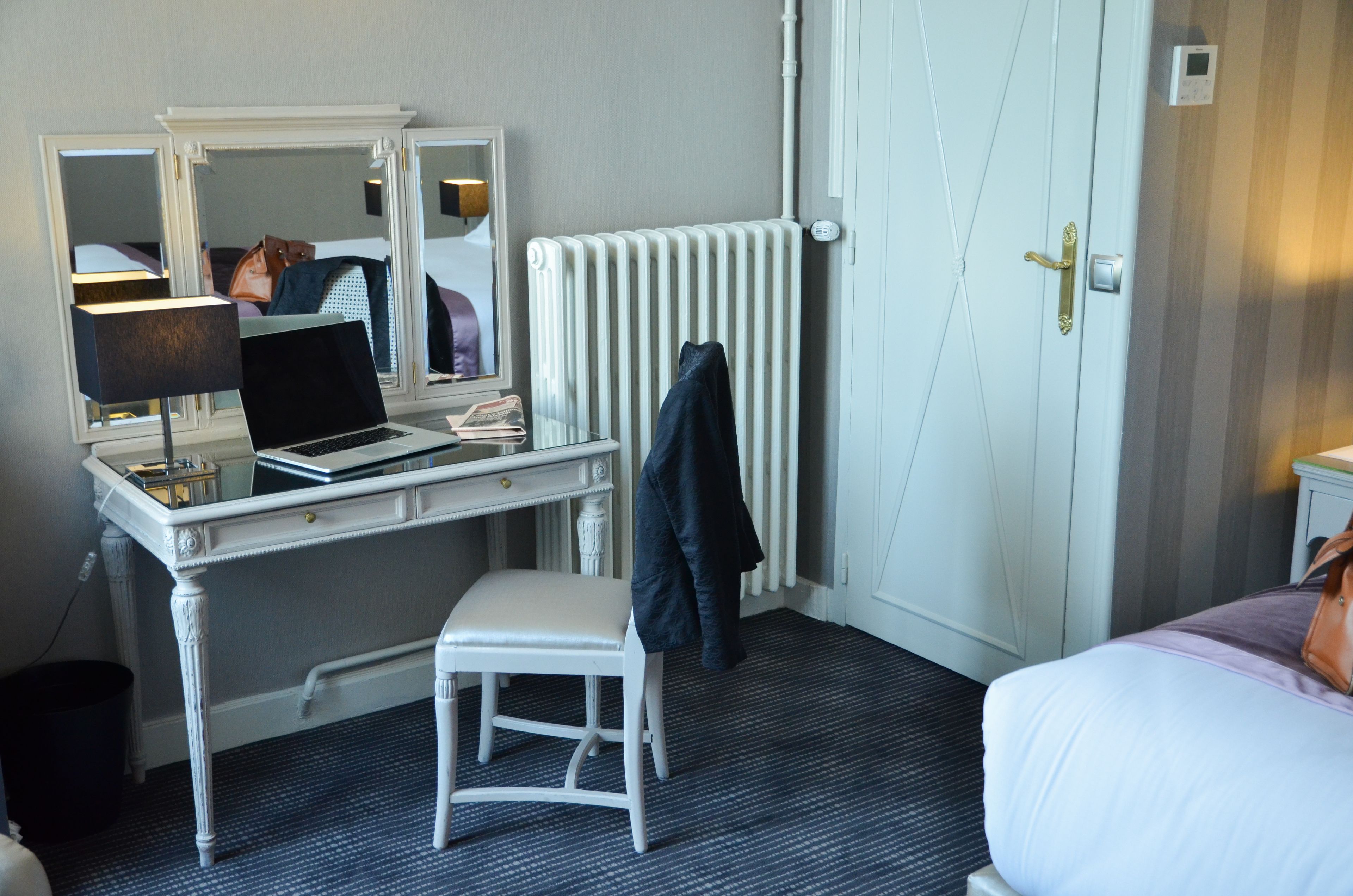 A hotel room with a white desk, a laptop, a mirror, a radiator, and a bed with a purple blanket.