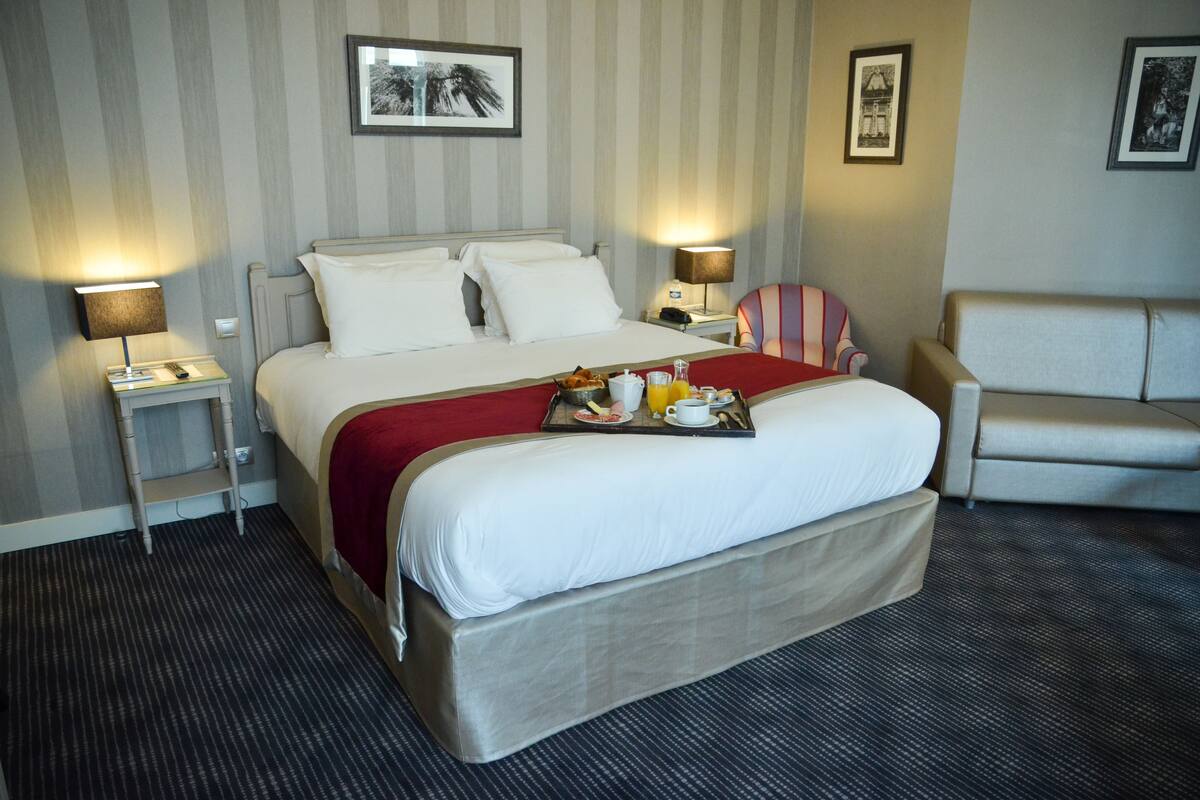 Habitación de hotel con una cama grande, dos mesitas de noche, una manta roja, una bandeja con comida para el desayuno, una pared a rayas y dos cuadros enmarcados.