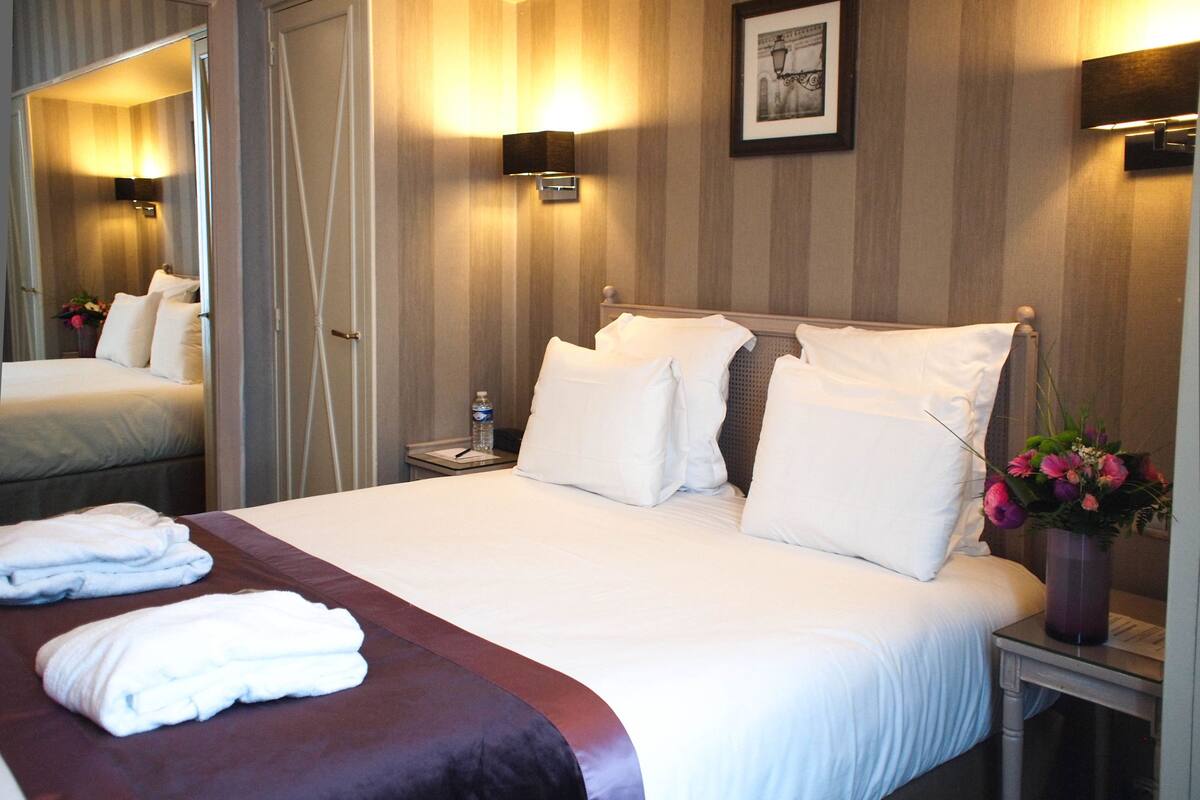 Habitación de hotel con una cama grande, una mesita de noche con lámpara, un espejo y un cuadro en la pared.
