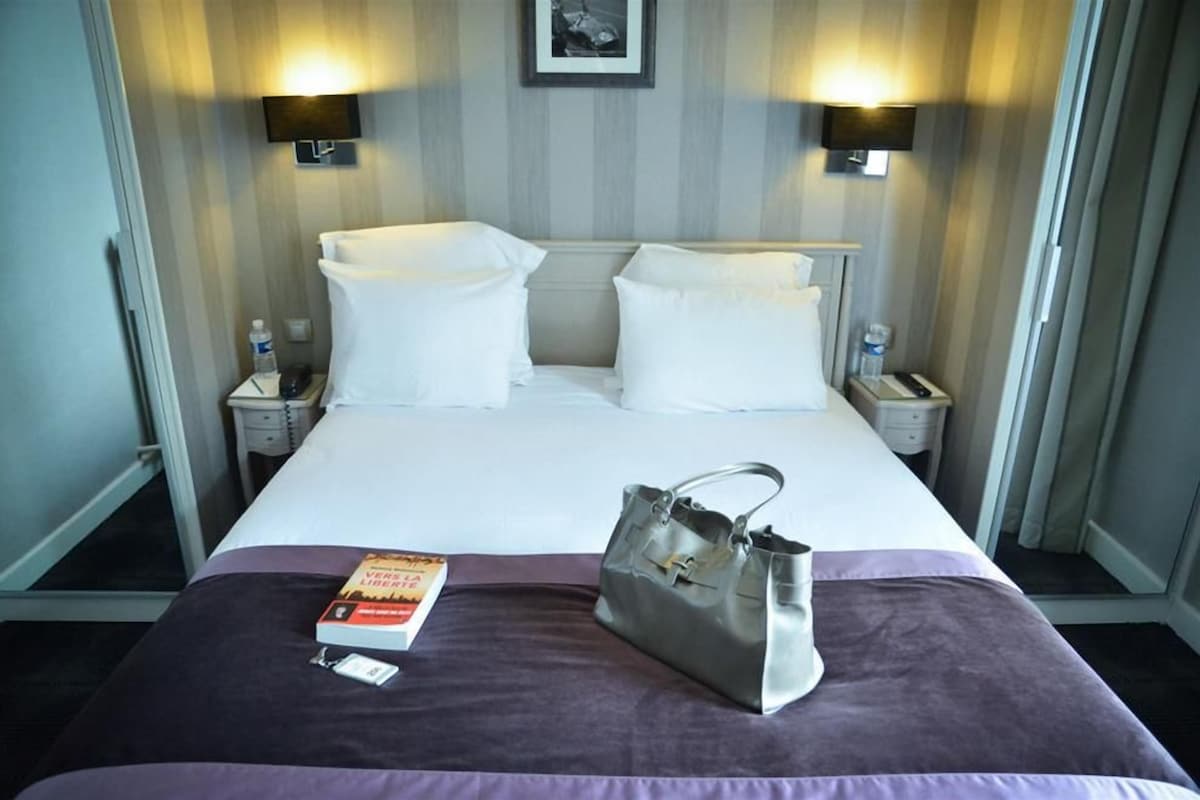 A hotel room with a bed, two bedside tables, a nightstand, a lamp, a book, and a handbag.