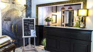 Un espace d’accueil d’hôtel avec un comptoir noir, un grand miroir et un tableau noir servant de menu.