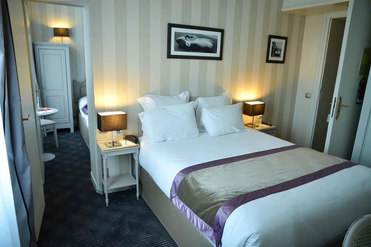 A hotel room with a large bed, two bedside tables with lamps, a small desk with a chair, a framed picture on the wall, and a door to the right.