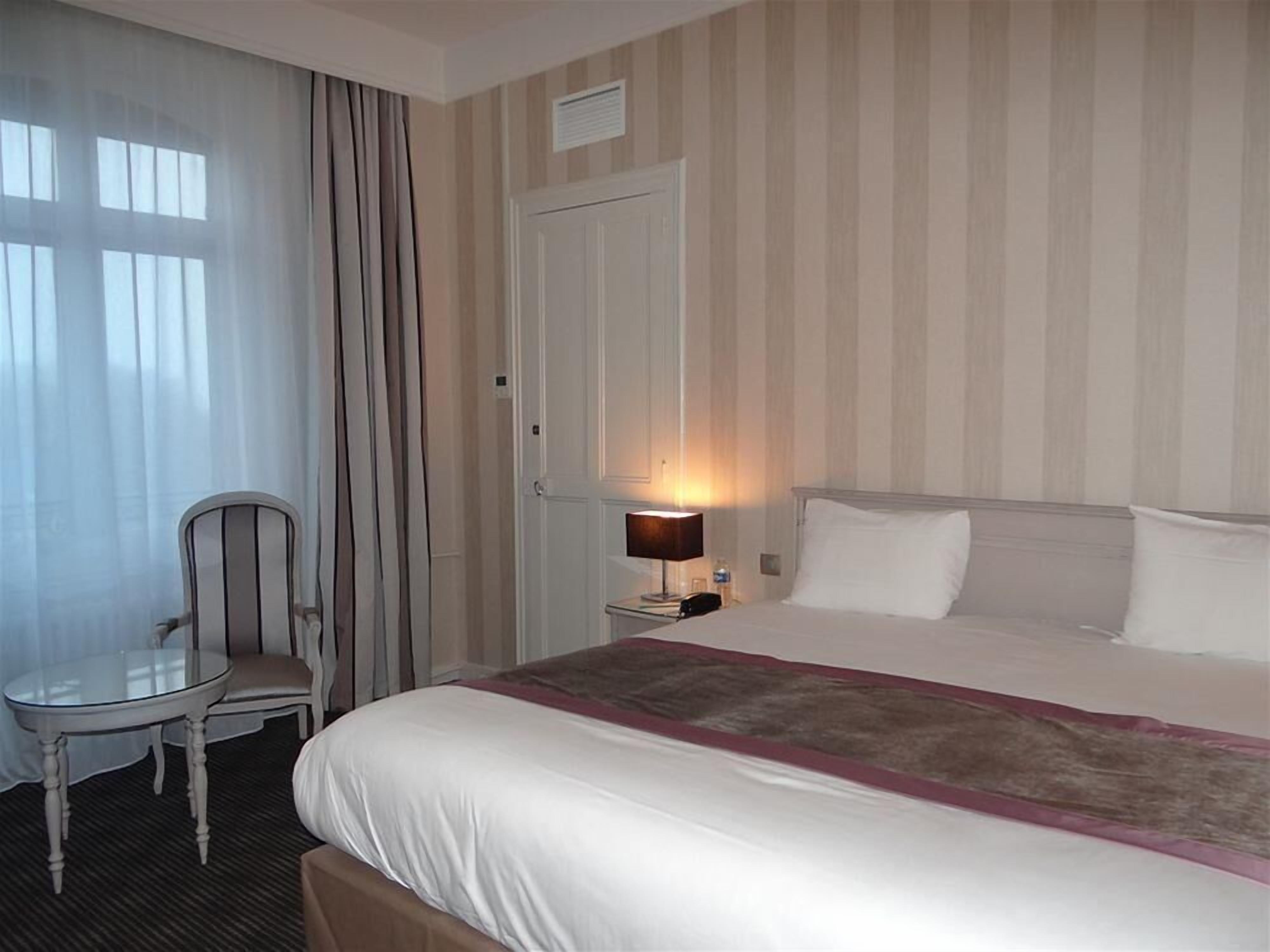 Una habitación de hotel con una cama, una silla, una mesita de noche con lámpara y una puerta.