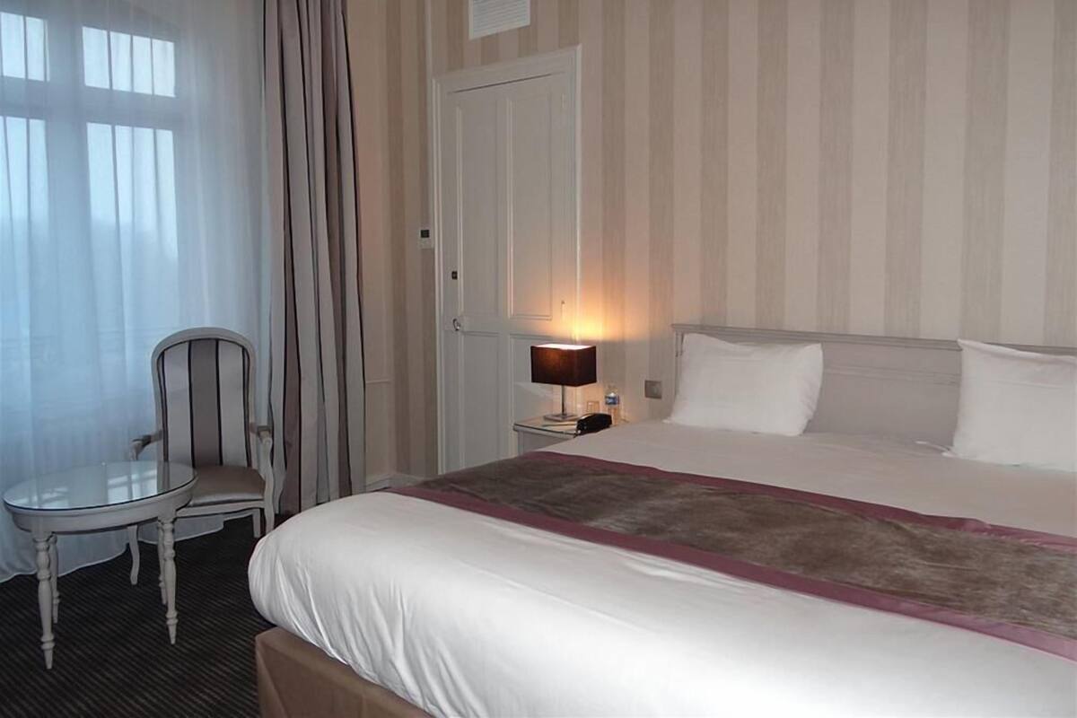 Habitación de hotel con una cama, una silla, una mesita de noche con lámpara y una puerta.