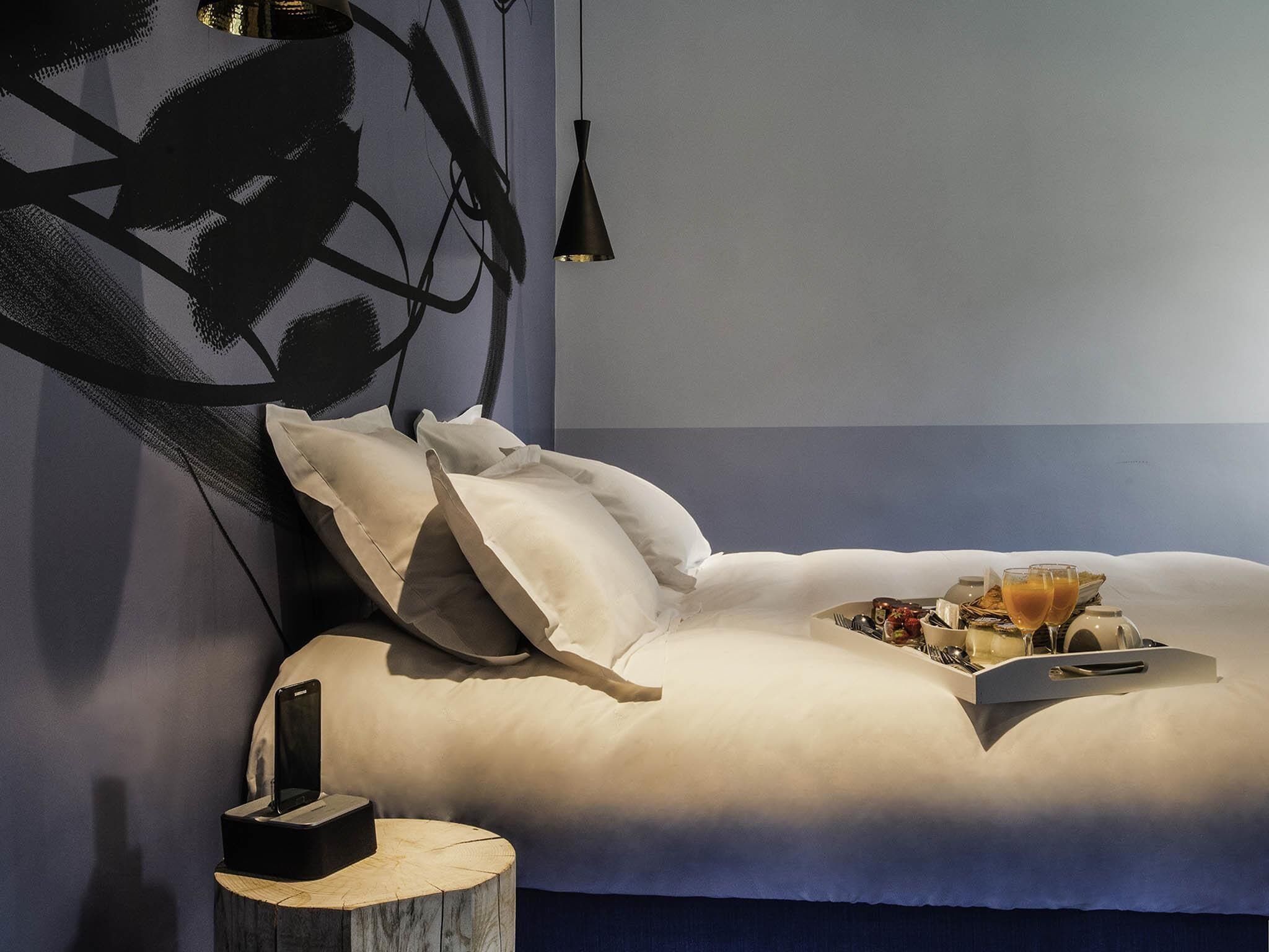 A modern bedroom with a neatly made bed, a bedside table with a lamp, and a tray with breakfast items.