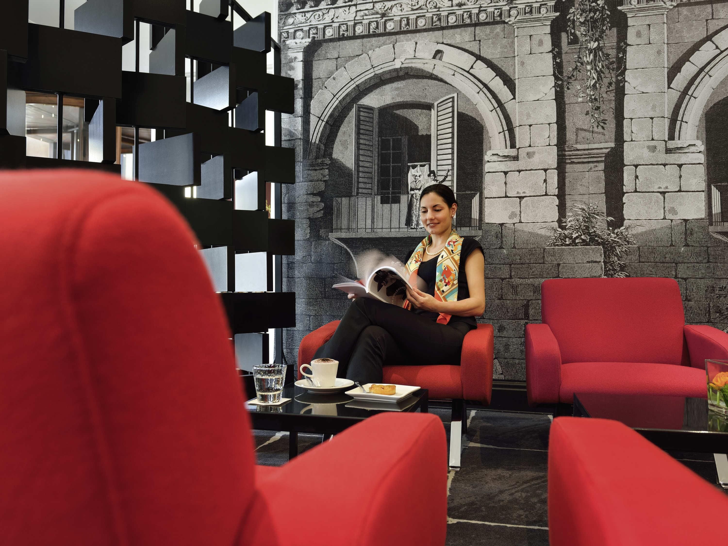 A woman sitting on a red chair reading a book in a room with a black and white patterned wall and a stone archway.