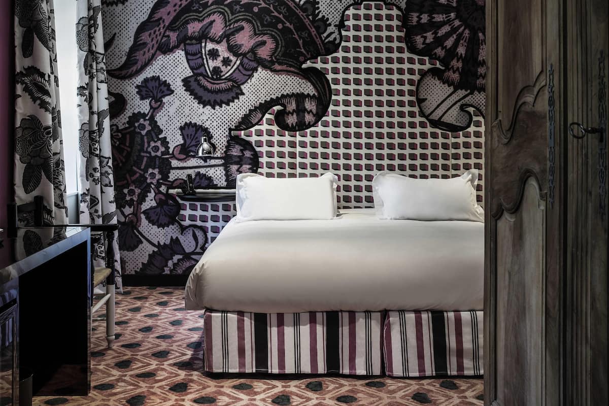 A bedroom with a patterned wall, a bed with white bedding, a striped ottoman, and a nightstand with a lamp.