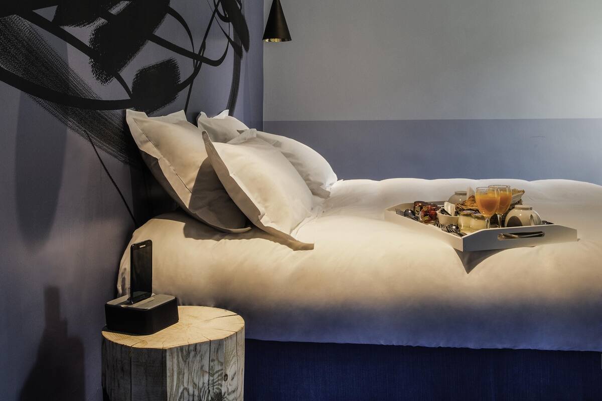 A bedroom with a bed, pillows, a bedside table with a phone, and a tray with food and drinks.