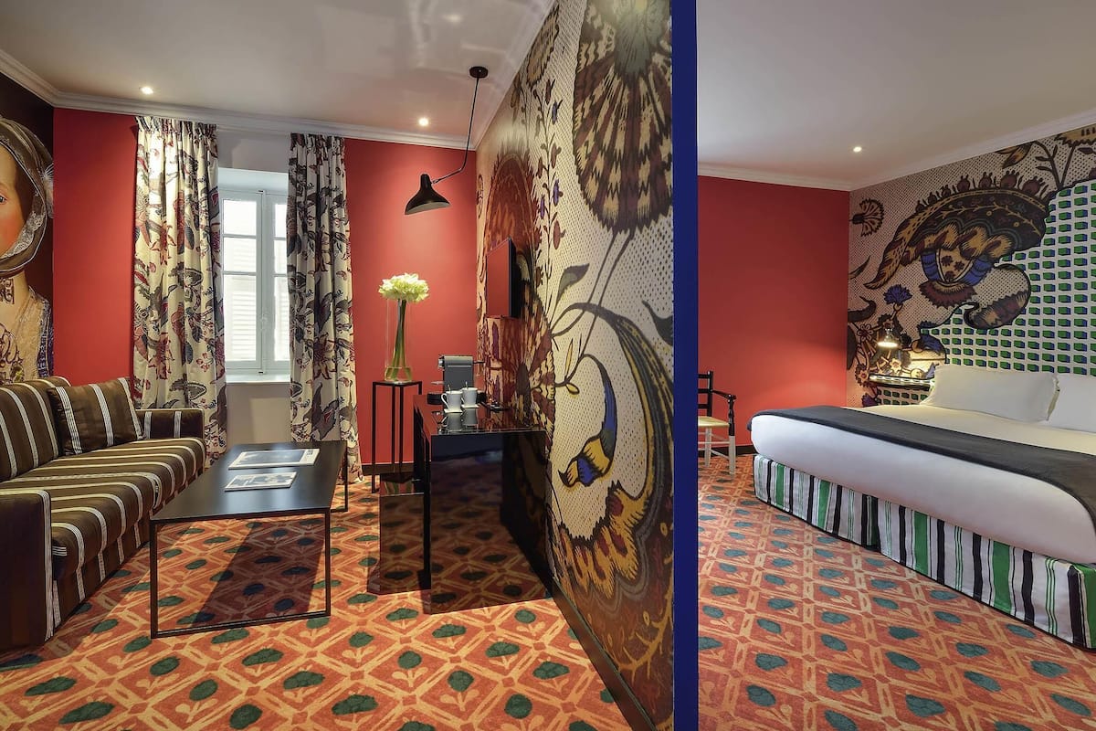 A hotel room with a sofa, a dining table, a decorative vase, a patterned wall, and a bed with striped bedding.