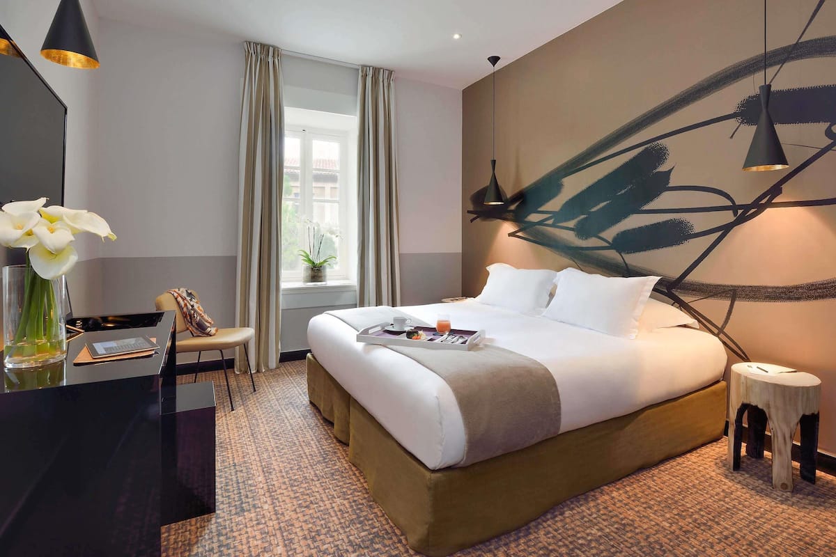 A hotel room with a bed, bedside table, lamp, window with curtains, and a wall art piece.