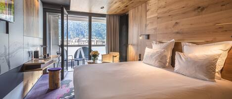A modern hotel room with a large bed, wooden panel walls, a desk with a chair, and a view of a mountainous landscape through large windows.
