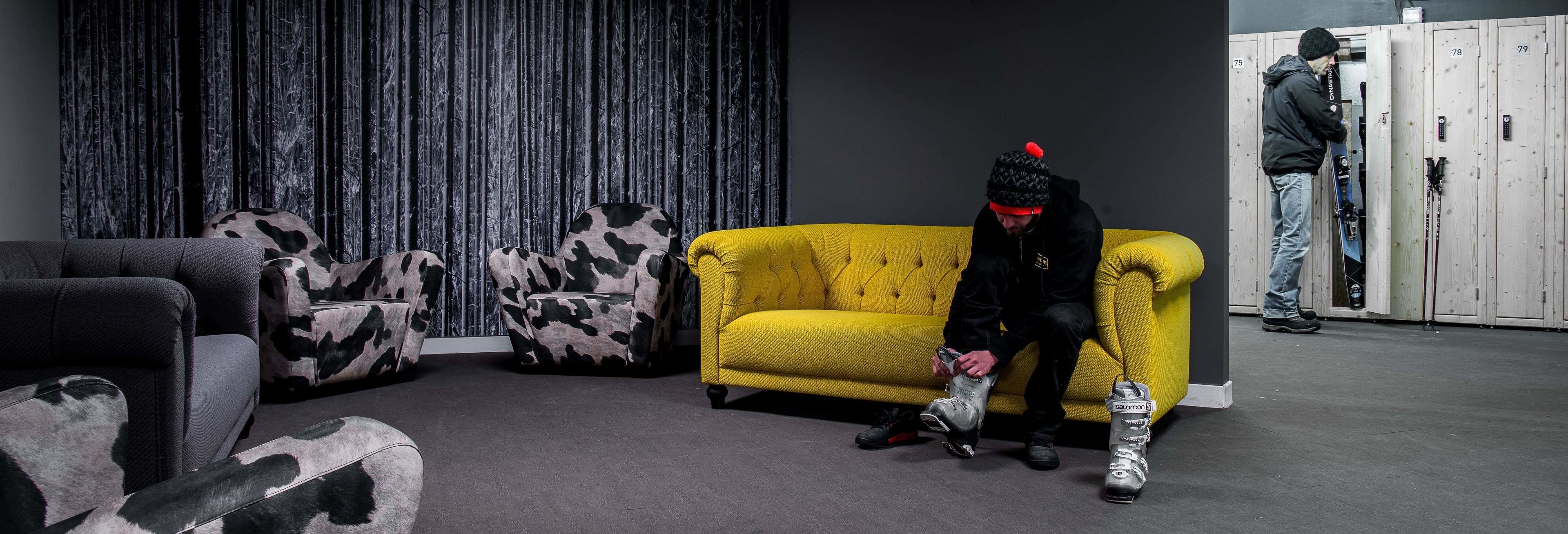A room with a yellow sofa, a patterned armchair, and a black and white patterned chair. There is a person sitting on the sofa, and another person standing near a set of lockers.