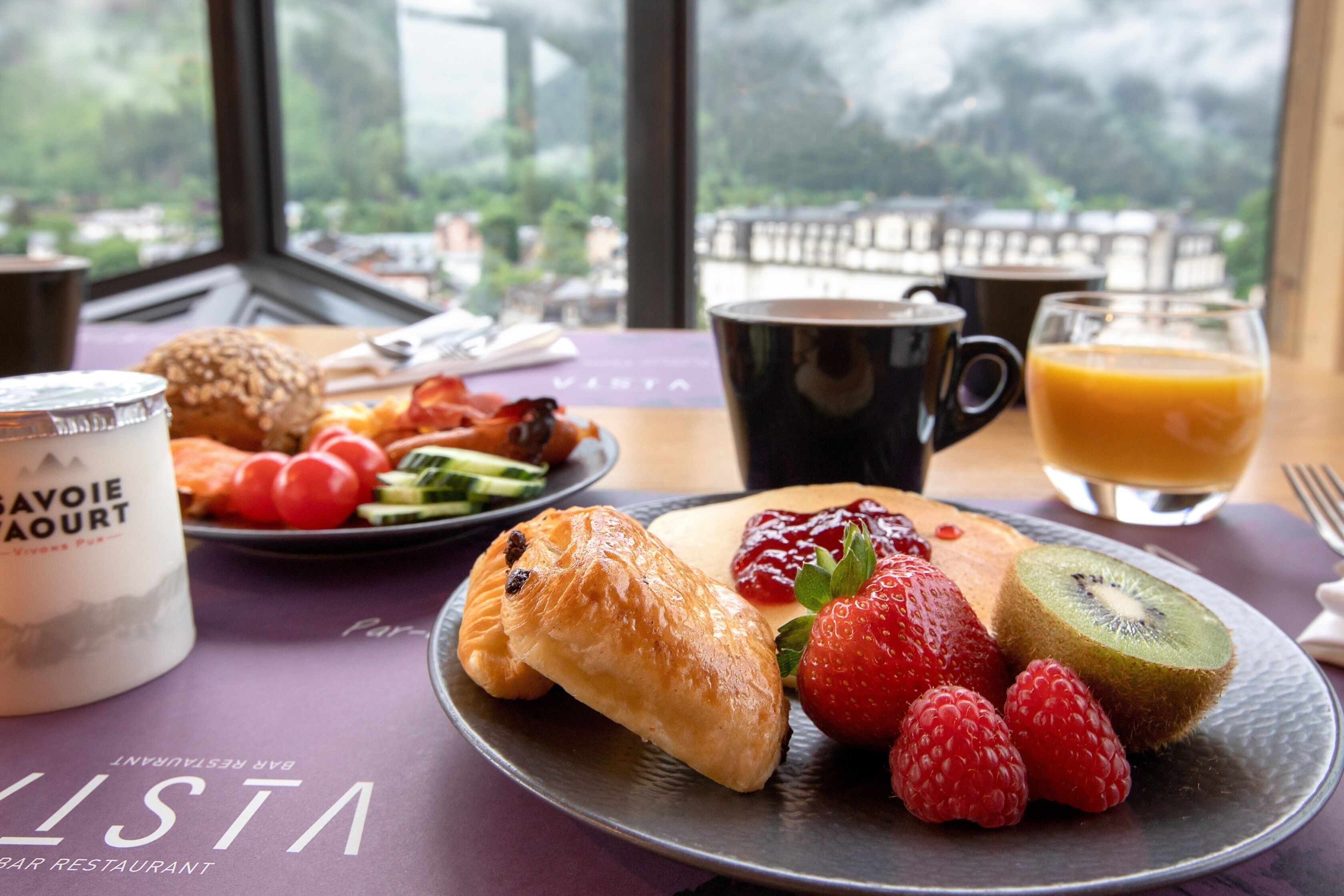 Una mesa de desayuno con cruasanes, fresas, frambuesas, kiwi y un vaso de jugo de naranja.