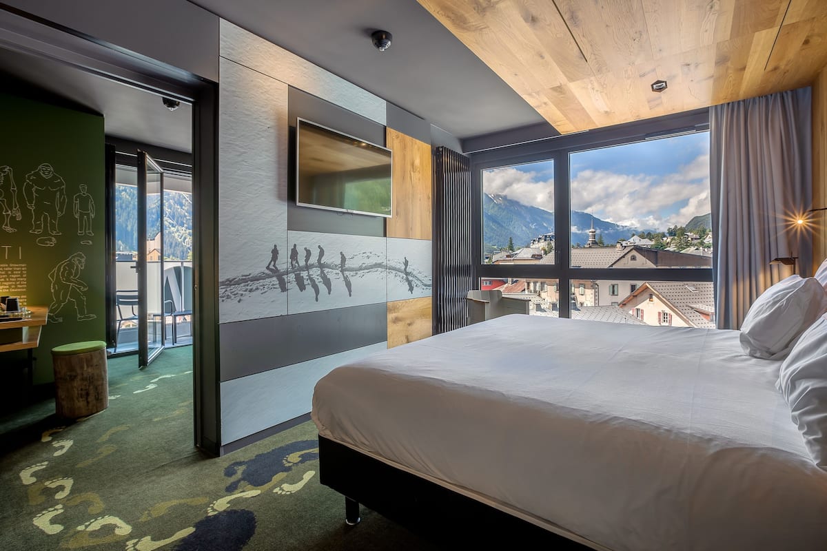 A modern hotel room with a large bed, a TV, and a view of mountains.