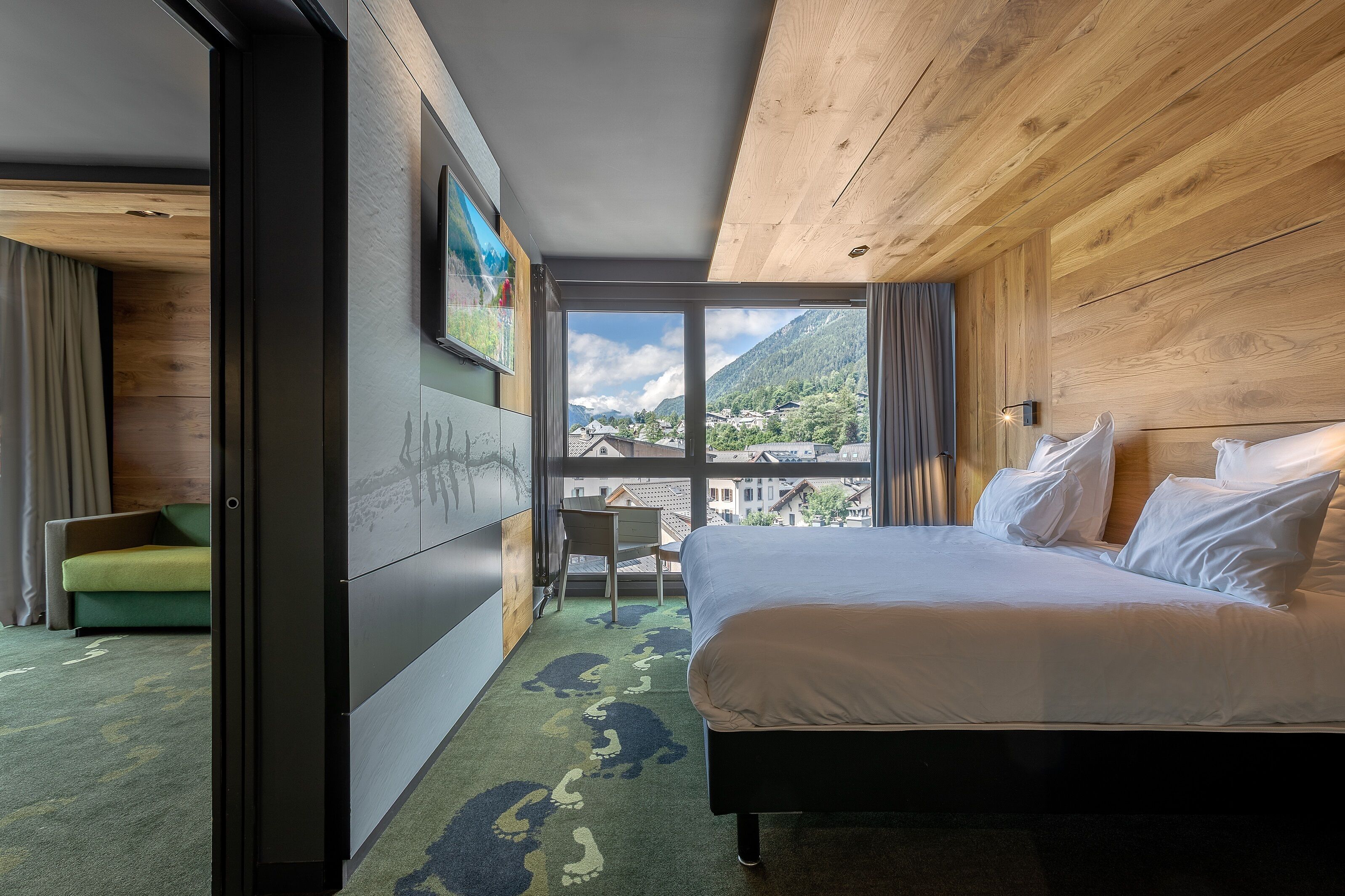 A hotel room with a large bed, a view of a mountain village, and a green sofa.