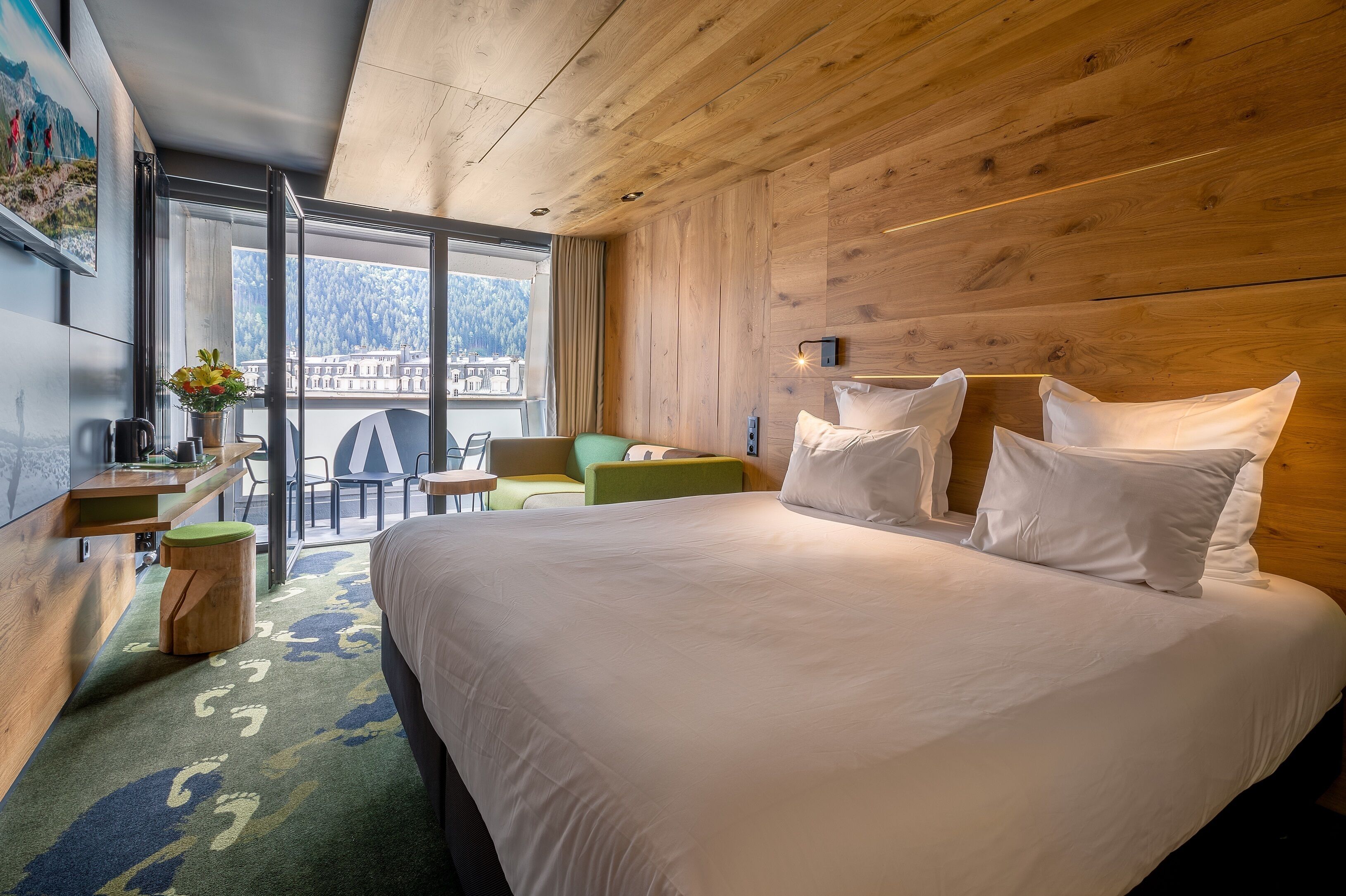 Habitación de hotel con una cama grande, un escritorio con silla, una mesita y vistas a una montaña y a la ciudad a través de un ventanal amplio.