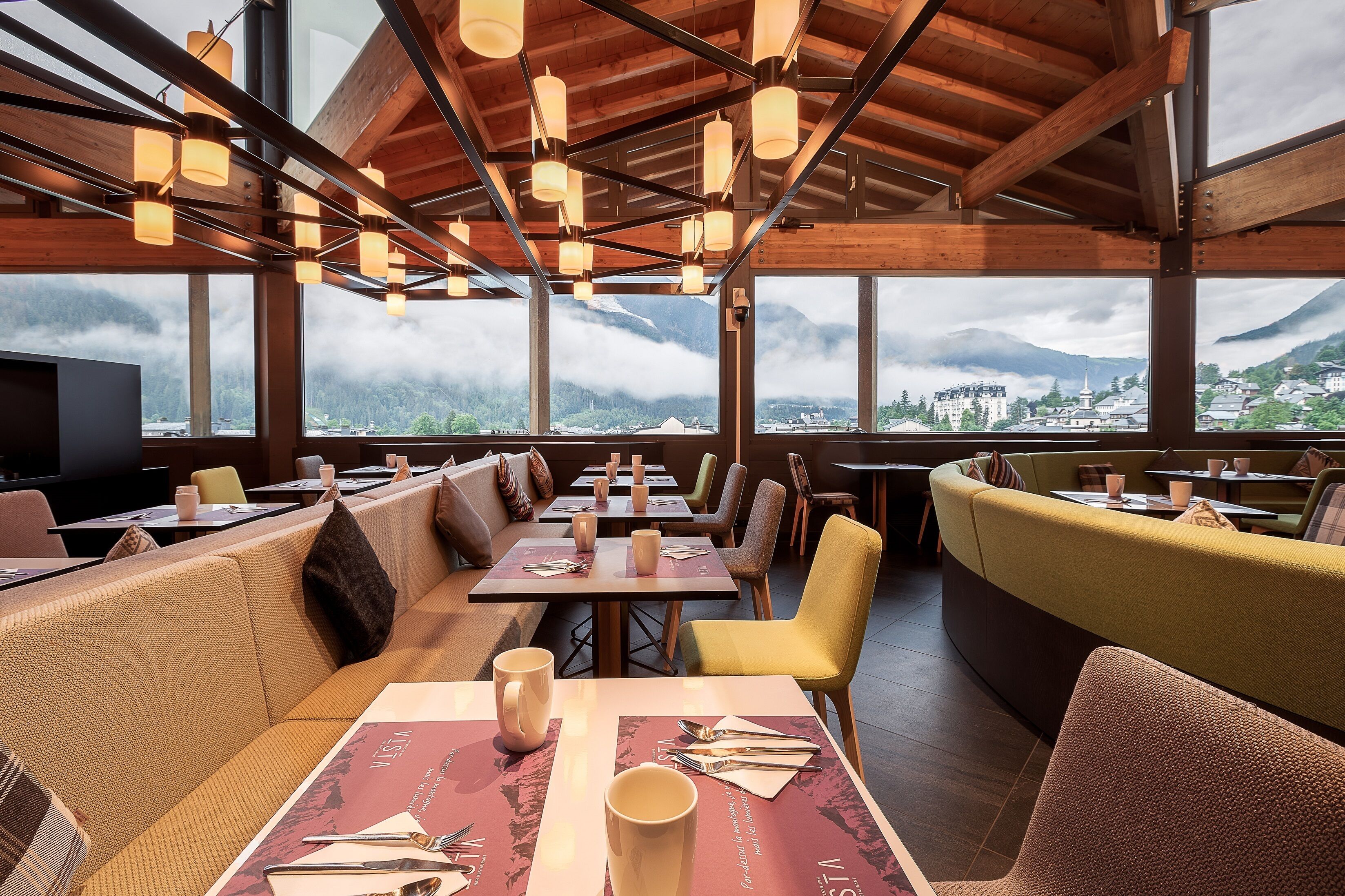 Un restaurante con vigas de madera, ventanas amplias y vistas a las montañas. Las mesas están preparadas con cubiertos y servilletas.