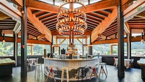 A restaurant with a central bar, wooden ceiling beams, and large windows offering a mountain view. - Alpina Eclectic Hotel (Chamonix-Mont-Blanc)