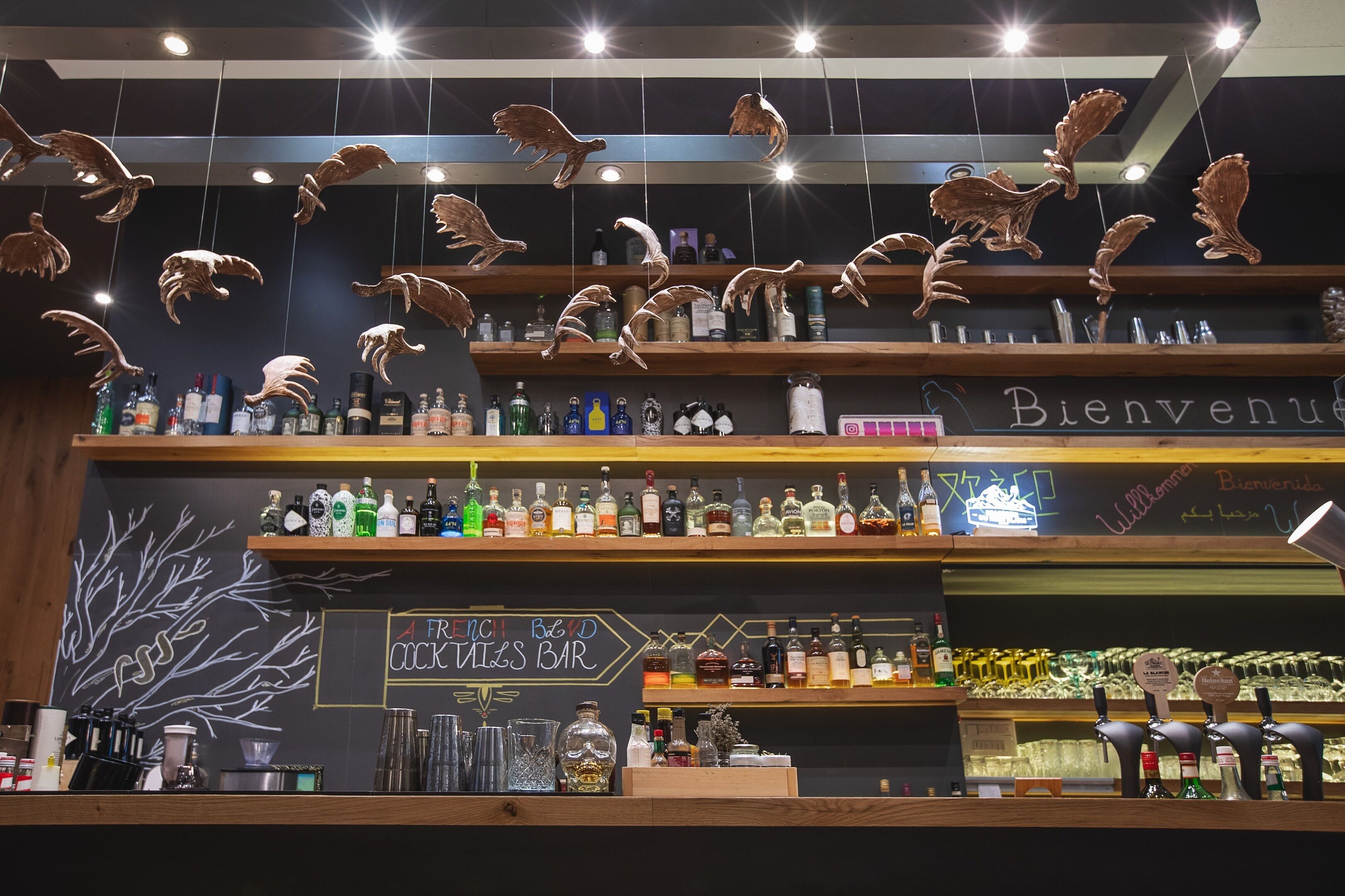Un bar con estantes de madera, adornos de pájaros colgantes y un cartel de pizarra que dice "FRENCH COCKTAILS BAR".