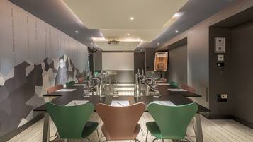 A conference room with a long table, green and brown chairs, a projector screen, and a wall with geometric patterns and text.