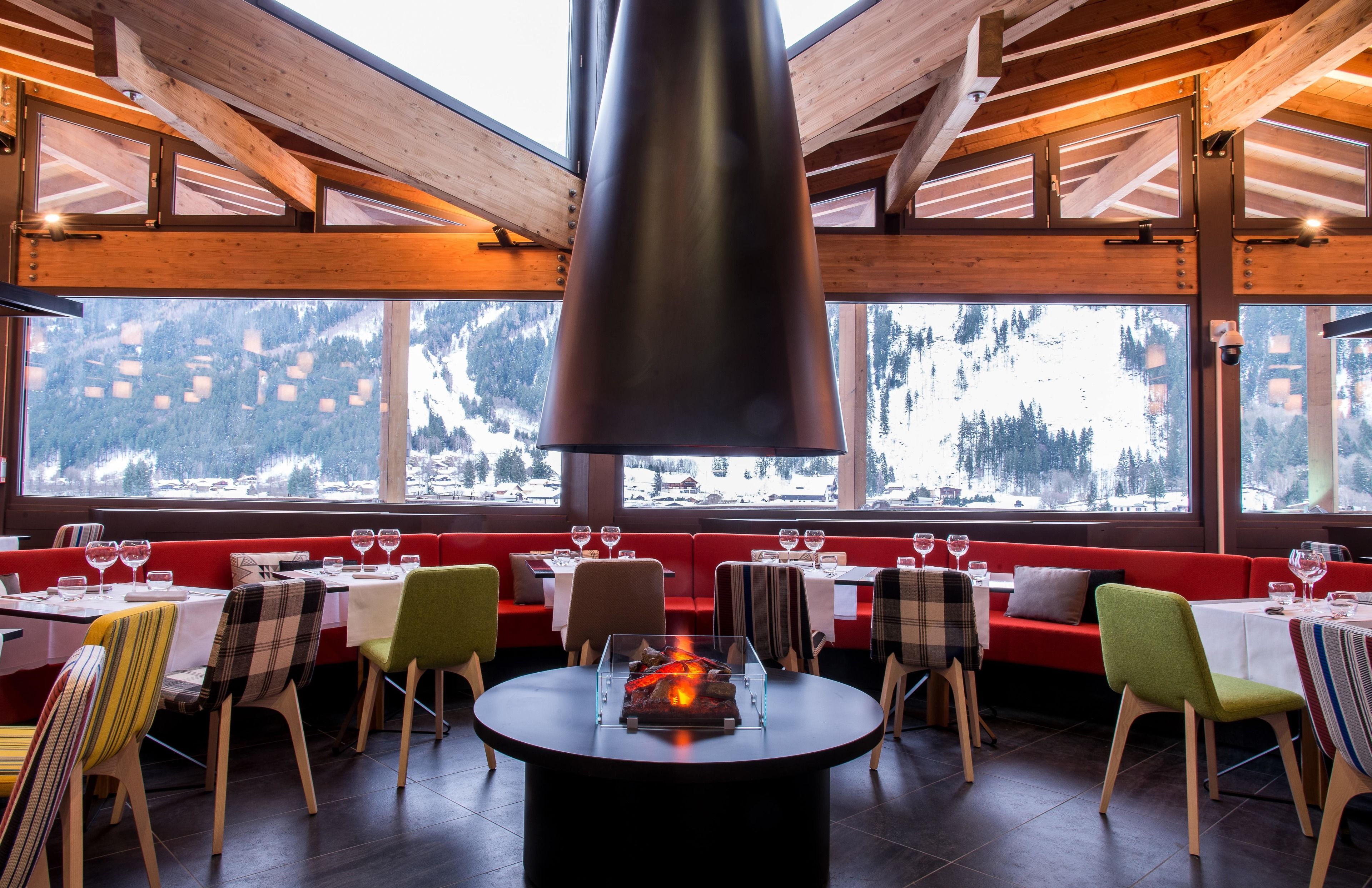 Ein Restaurant mit Holzbalken, großen Fenstern mit Blick auf einen verschneiten Berg, roter Sitzgelegenheit und einem zentralen Tisch mit Feuerstelle.