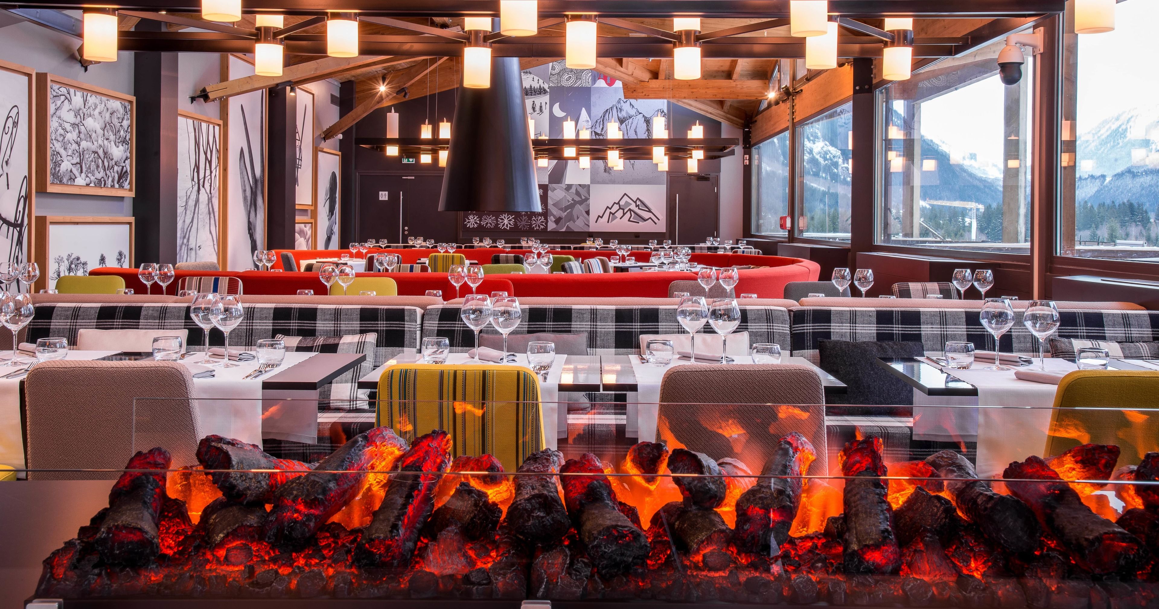 A restaurant with a long communal dining table, individual tables, and a fireplace with burning wood.