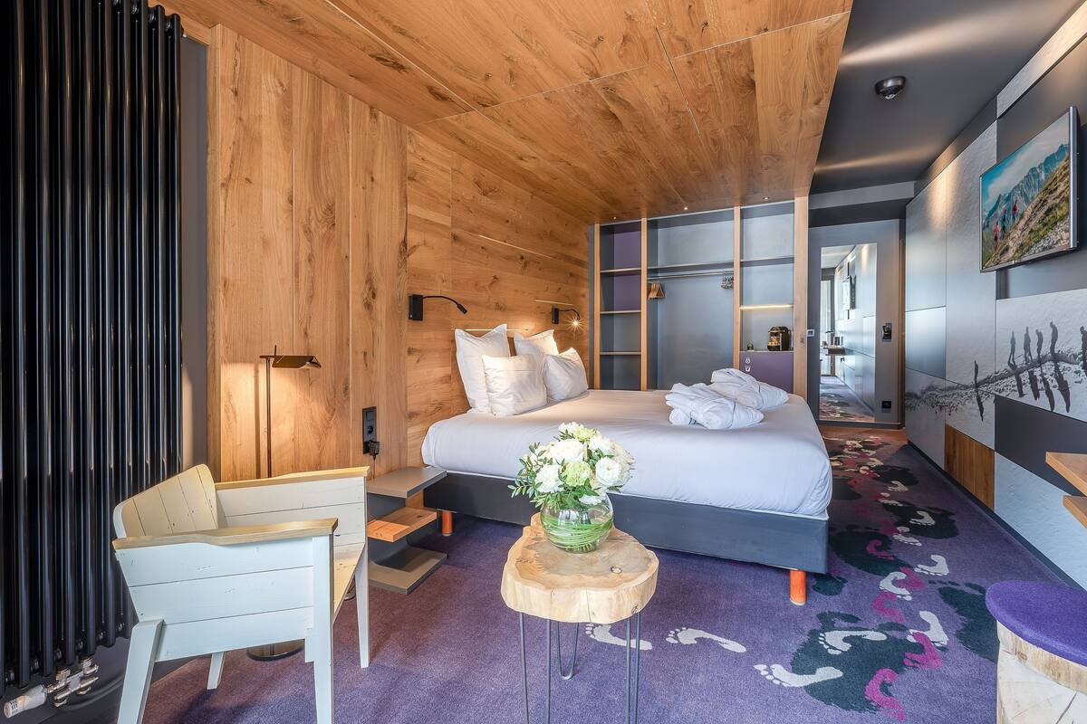 A modern bedroom with a wooden ceiling, a large bed with white linens, a small wooden side table with a vase of flowers, and a purple carpet with a mountain design.
