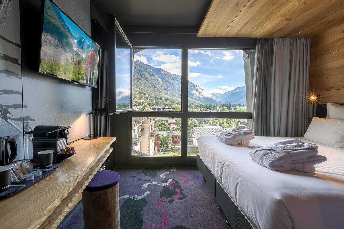 A hotel room with a large window offering a mountain view, a bed with neatly folded towels, a desk with a coffee maker, and a mounted TV.