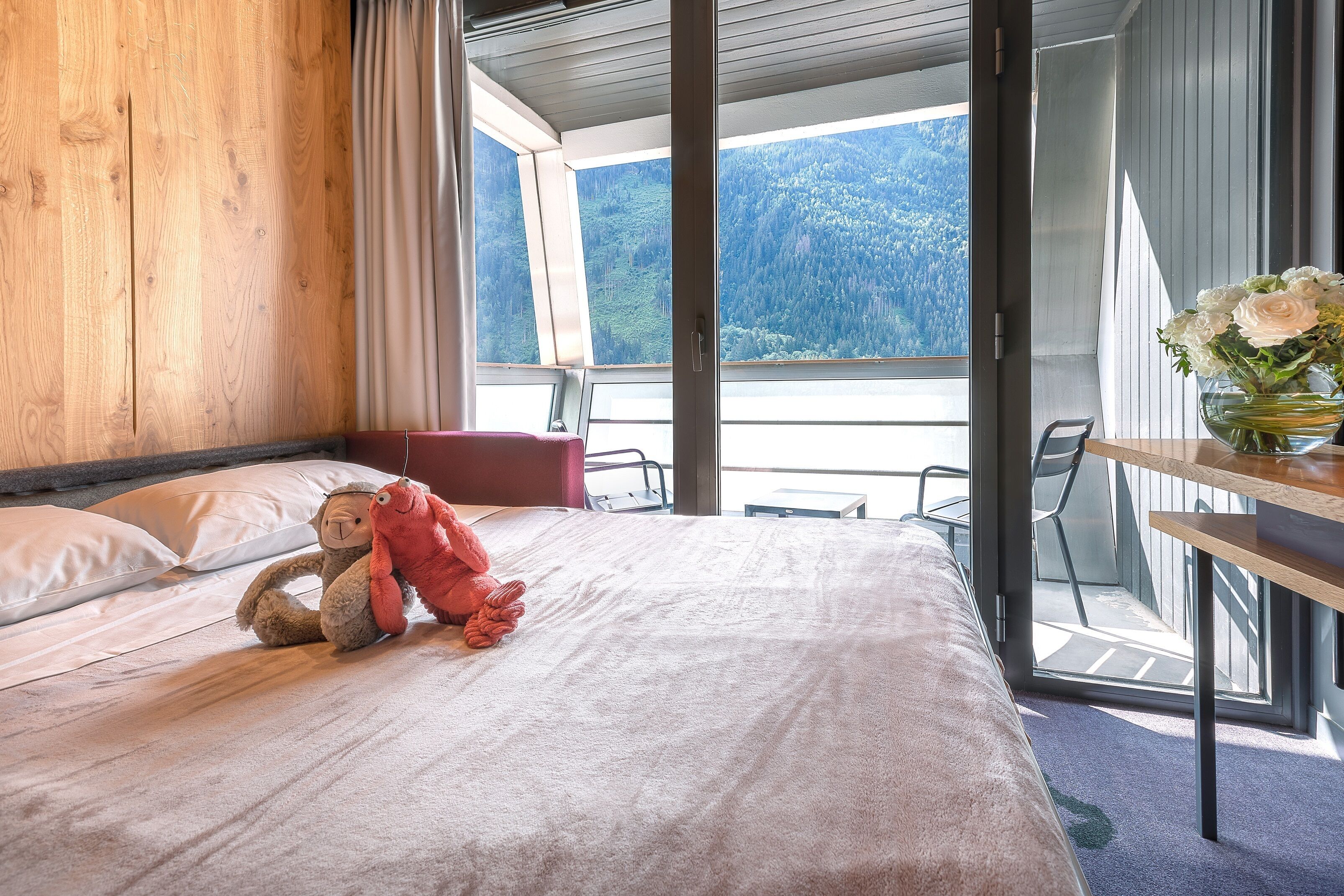 A bedroom with a bed, a plush toy, a vase with flowers, and a view of a forest through a large window.